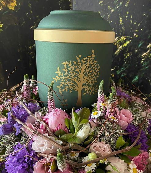 Grüne Urne mit goldenem Baum-Motiv, umgeben von einem Blumenkranz in Rosa, Lila und Weiß, liebevoll arrangiert von Ihrem Bestatter in Berlin für eine Naturbestattung.