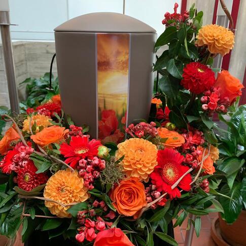 Dezente Urne mit Sonnenuntergang-Motiv, umgeben von leuchtendem Blumenkranz aus Rosen, Gerbera und Dahlien in Orange und Rot, stilvoll arrangiert von Ihrem Bestatter in Berlin.
