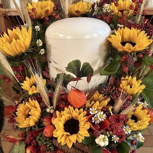 Weiße Urne, umgeben von Sonnenblumen, Herbstblumen und Ähren.