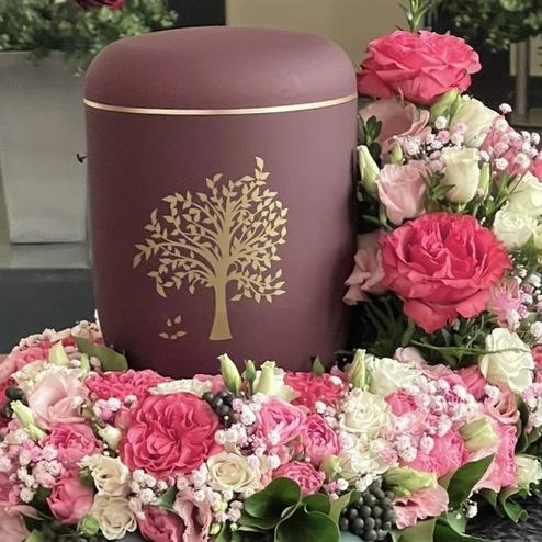 Bordeauxfarbene Urne mit goldener Baumgravur, umgeben von Rosenschmuck in Rosa und Weiß.