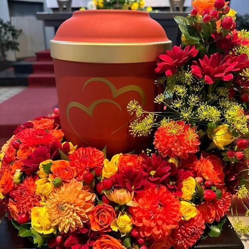 Orangefarbene Urne mit goldenen Herzen, umgeben von leuchtendem Blumenschmuck in Orange, Rot und Gelb.