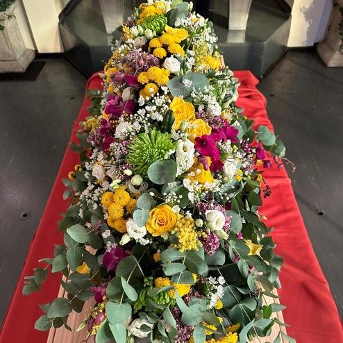 Farbiges Blumengesteck mit gelben Rosen und bunten Blumen schmückt den Sarg bei einer Trauerfeier, liebevoll arrangiert vom erfahrenen Bestatter in Berlin.