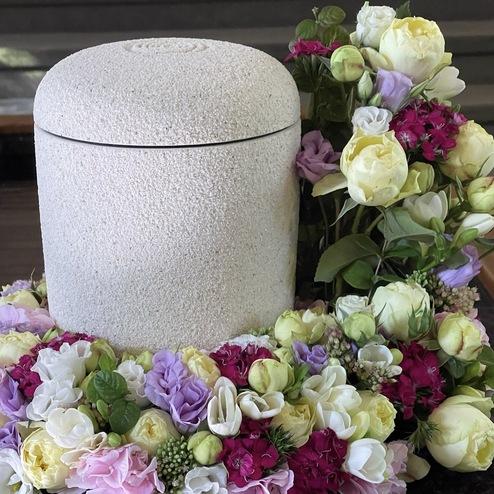 Schlichte, helle Urne, umgeben von einem farbenfrohen Blumenarrangement mit Rosen und Lisianthus, stilvoll präsentiert von Ihrem Bestatter in Berlin.