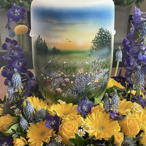 Handbemalte Urne mit Landschaftsmotiv, umgeben von gelben Rosen, Gerbera und blauen Blüten, liebevoll gestaltet von Ihrem Bestatter in Berlin für eine persönliche Abschiednahme.