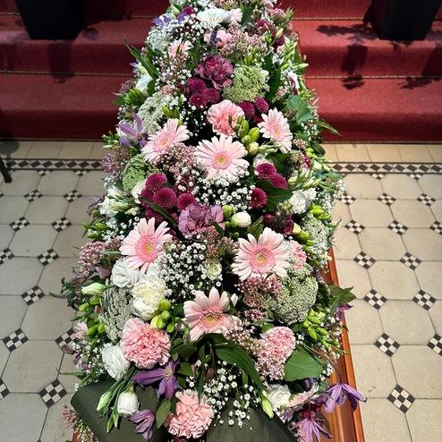 Elegantes Blumengesteck mit Gerbera, Nelken und Schleierkraut in Rosa und Lila auf einem Sarg, stilvoll arrangiert von Ihrem Bestatter in Berlin für eine persönliche Trauerfeier.