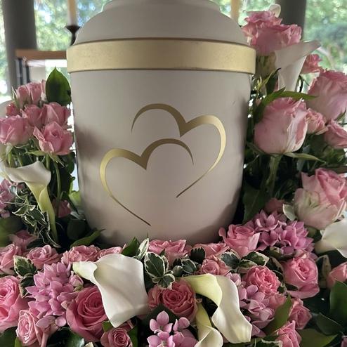 Weiße Urne mit goldenen Herzen, umgeben von rosa Rosen, Calla und Hortensien, stilvoll arrangiert von Ihrem Bestatter in Berlin für eine liebevolle Abschiednahme.