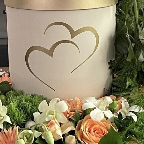 Weiße Urne mit goldenen Herzen, umgeben von einem zarten Blumenkranz mit Rosen und Gerbera in Apricot und Weiß, arrangiert von Ihrem Bestatter in Berlin für eine persönliche Abschiednahme.