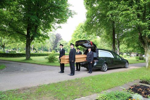 Vier Trauernde in Anzügen stehen am Straßenrand, während ein Sarg aus einem schwarzen Bestattungsfahrzeug entnommen wird.