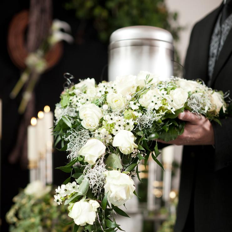 Nahaufnahme eines Blumenkranzes mit weißen Rosen, gehalten von einer Person vor einem silbernen Urnengrab.