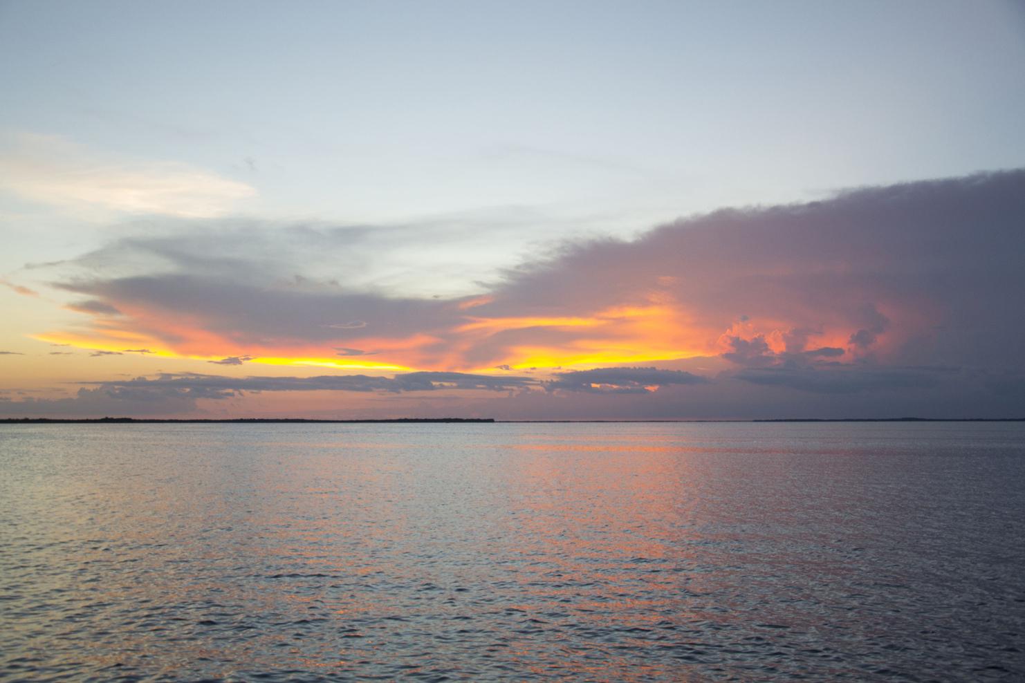 Blick auf einen ruhigen See mit orangefarbenem Sonnenuntergang und sanften Wolken am Horizont.