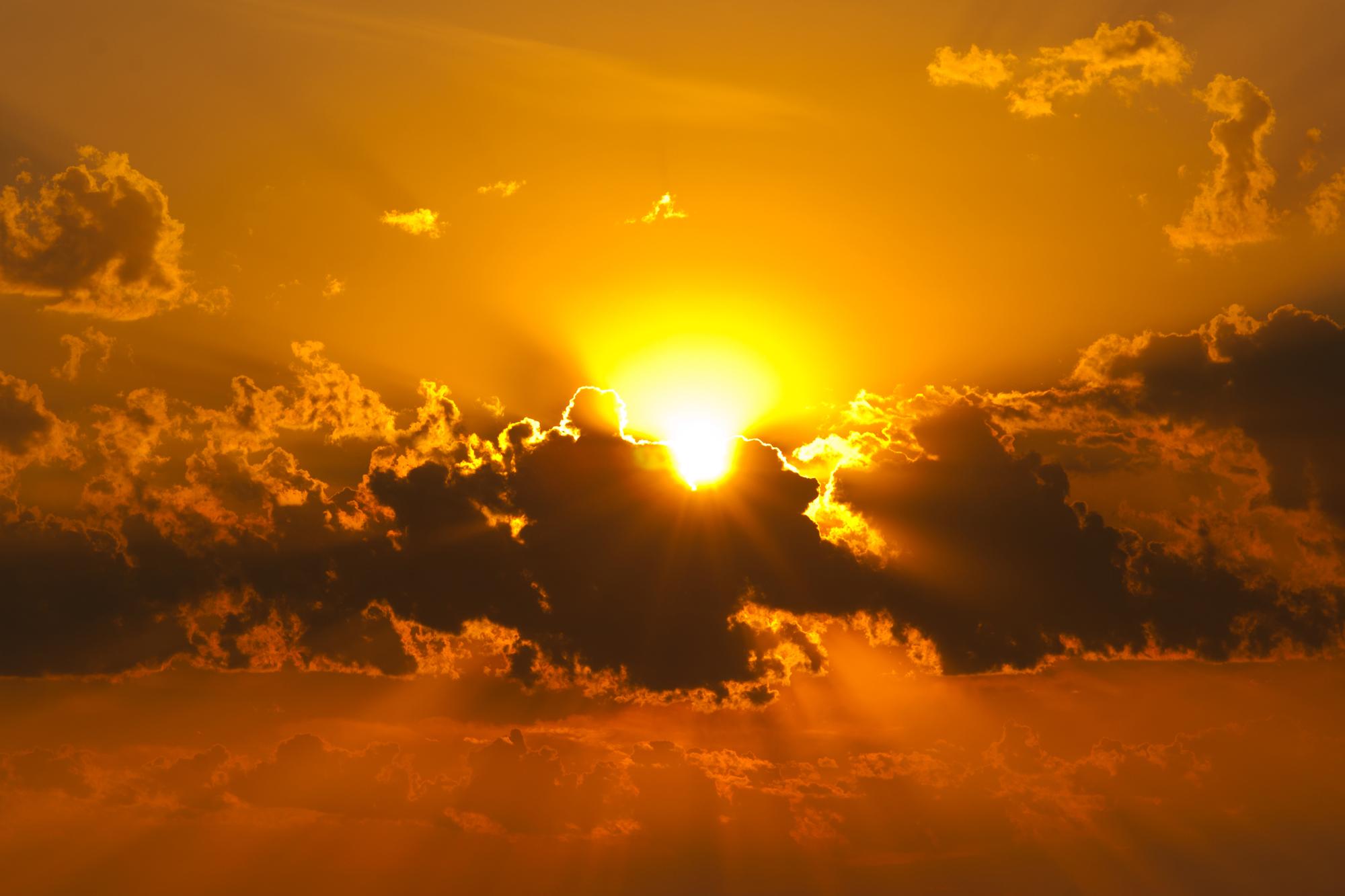 Sonnenuntergang mit goldener Sonne, die hinter dunklen Wolken strahlt, umgeben von orangefarbenem Himmel.
