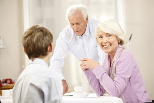 Lächelnde ältere Frau sitzt am Tisch, während ein Mann im Hintergrund Getränke serviert, ein Junge schaut sie an.
